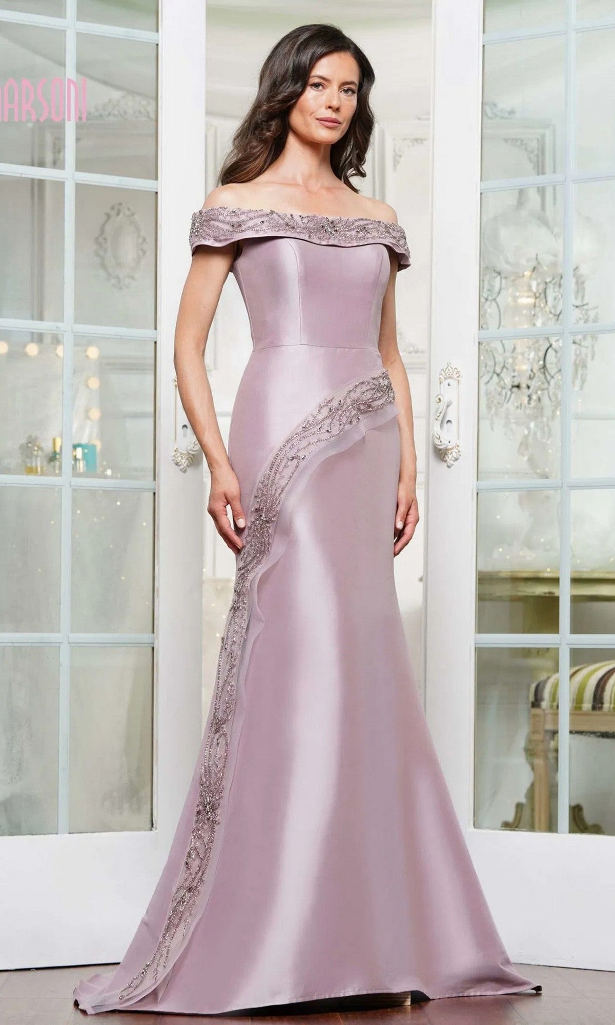 A woman in a dusty rose Marsoni MV1332 long formal dress with an off-shoulder, mermaid silhouette, lace and bead details stands indoors before glass doors.